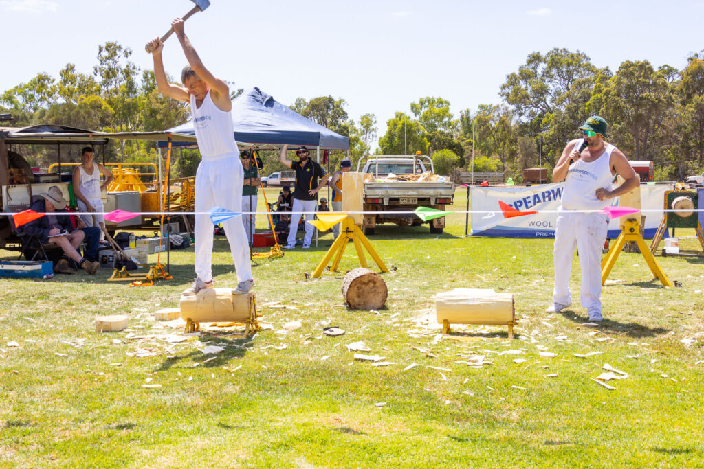 photo of a man dressed in white chopping the log he is standing on with an axe, with another man holding a microphone and commentating, at Darkan Sheepfest 2023.