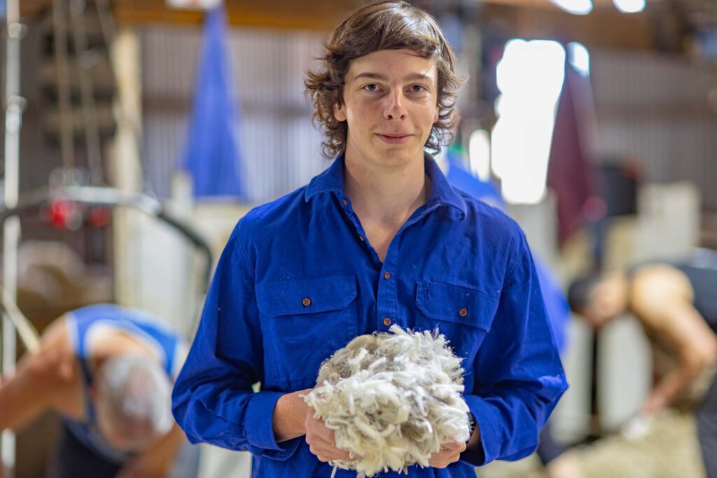 a teenaged boy wearing blue long-sleeved worksheet holds a sample of fine white merino wool in his hands as he looks at the camera. Behind him in the shearing shed, blurred in the background, shearers are bending down shearing sheep.