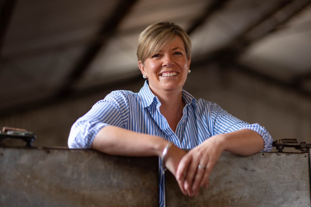 photo of smiling blonde woman wearing pearl stud earrings and blue and white striped shirt with arms resting on gate in a shearing shed. She has a broad smile and is looking at the camera.